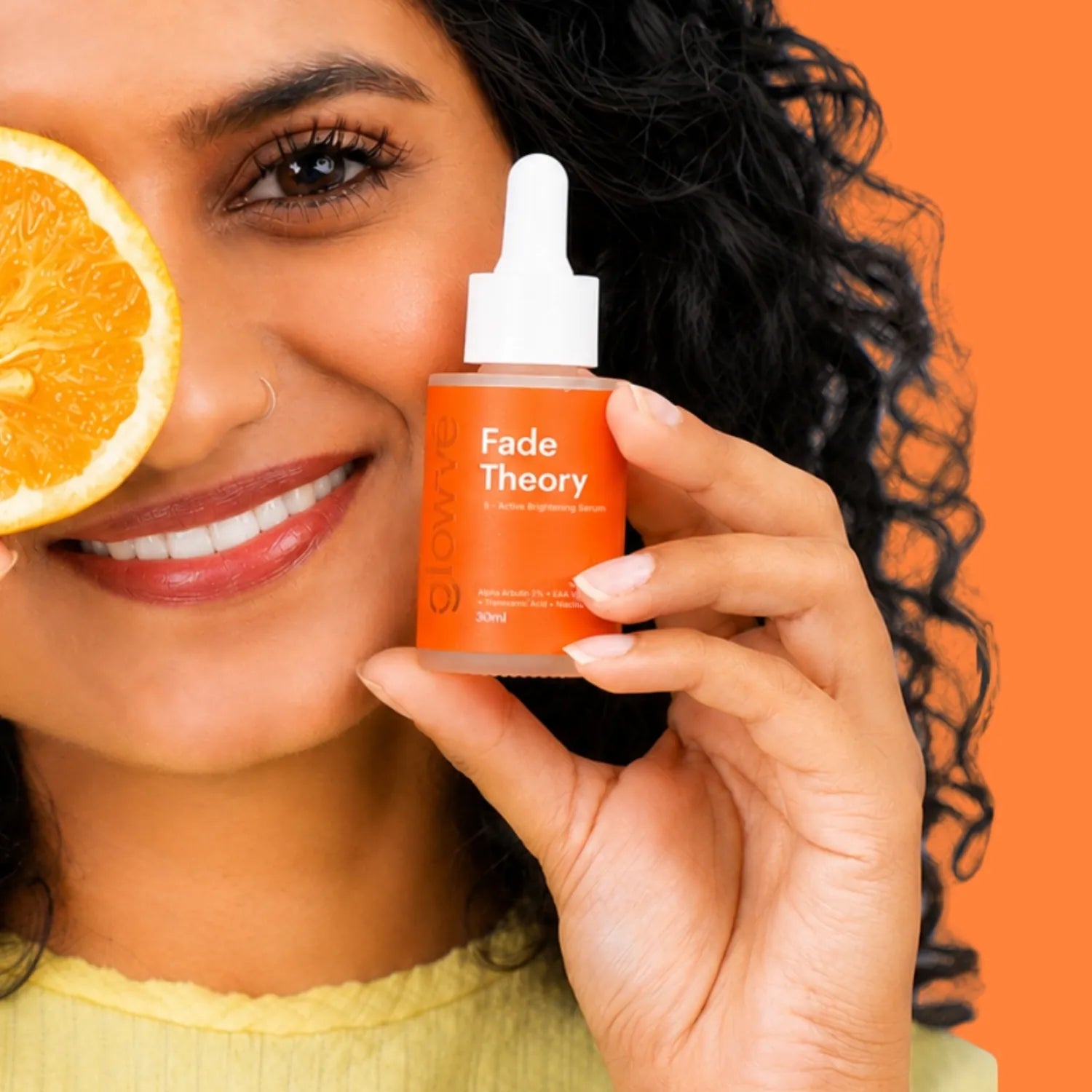 Woman holding Fade Theory 2% alpha arbutin serum beside her face, with a slice of orange, on an orange background